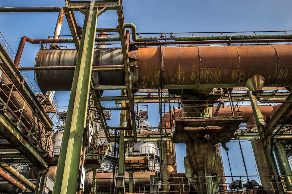 A low angle view of a rusty old industrial structure with pipes and beams against a clear blue sky.